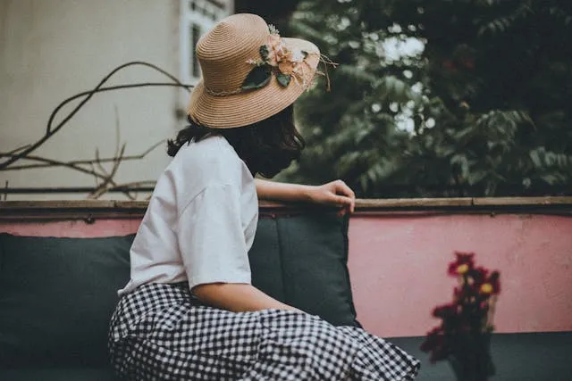 Image of woman sitting at a terrace looking out into the city and nature. The lady in the picture is wearing a white top with a black and white checkered long skirt. She is also wearing a beautiful hat which has floral decorations on it.