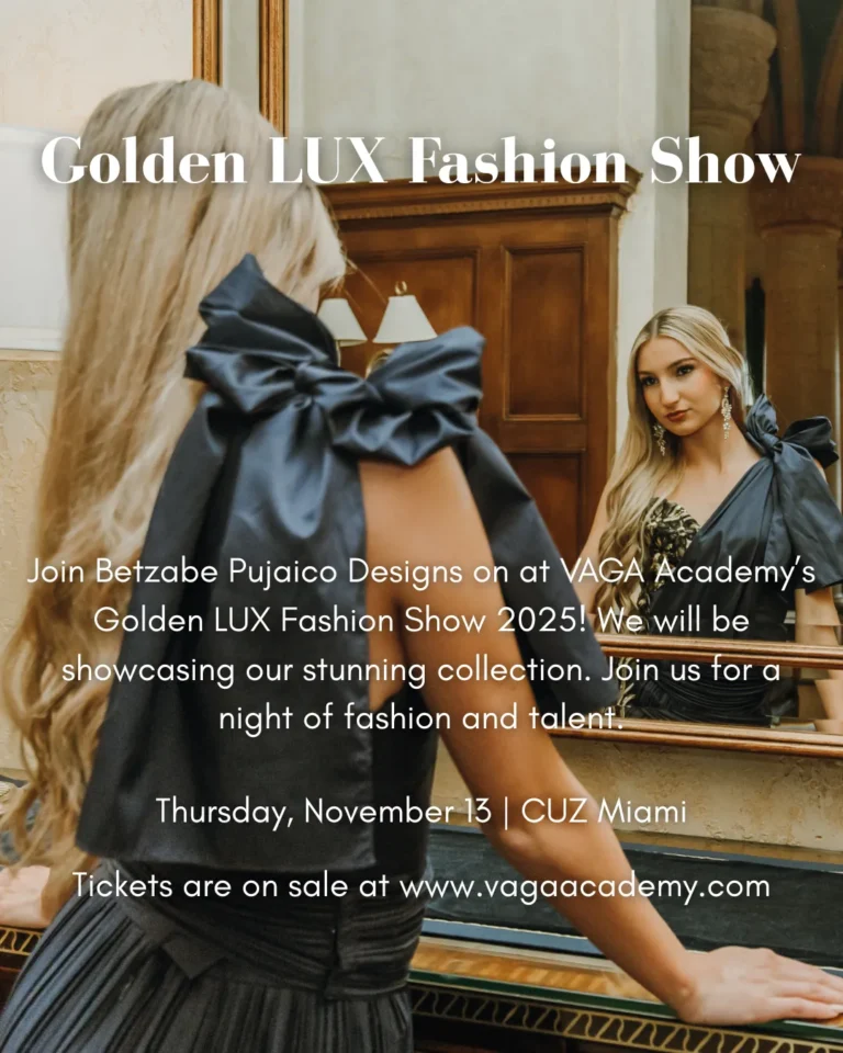 Image of woman standing in front of a mirror at building hall. She's wearing a luxurious grey dress which has a bow on her shoulder. The dress also has hints of gold. Her hair is blonde and she's wearing silver earrings. This picture was taken at the VAGA Academy's Golden LUX Fashion show in Miami Doral.